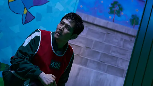 Gi-hun hält sein Knie und guckt mit einem wütenden Blick nach vorne. - Foto: No Ju-han/Netflix Gi-hun hält sein Knie und guckt mit einem wütenden Blick nach vorne. - Foto: No Ju-han/Netflix