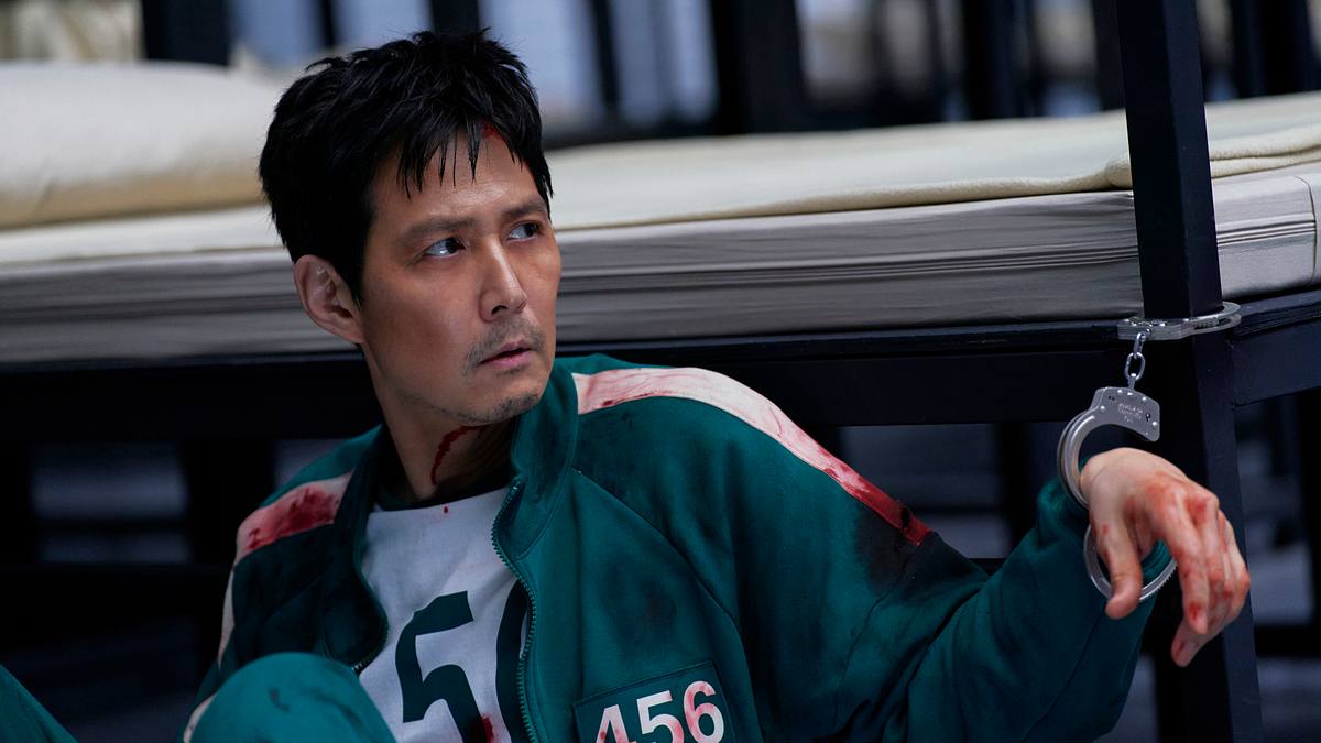 „Squid Game“ Lee Jung-jae Gi-hun ist mit Handschellen an sein Bett gekettet und schaut mit einem traurigem Bilck in die Ferne.