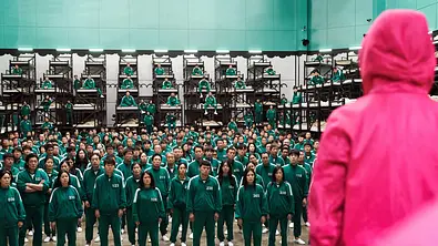Man sieht die Spieler und Spielerinnen der Serie „Squid Game“ in dunkelgrünen Trainingsanzügen. Vor ihnen steht eine der Wachen in einem pinken Trainingsanzug. - Foto: Netflix