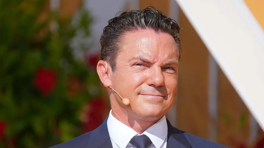 Stefan Mross Stefan Mross beim Ende von Immer wieder sonntags. Er trägt einen dunkelblauen Anzug mit Krawatte und trägt ein Headset-Mikrofon.