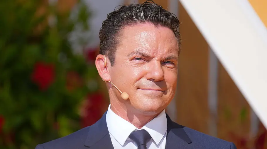Stefan Mross Stefan Mross beim Ende von Immer wieder sonntags. Er trägt einen dunkelblauen Anzug mit Krawatte und trägt ein Headset-Mikrofon.