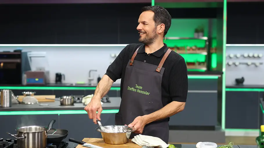 Steffen Henssler The Taste Steffen Henssler rührt lächelnd in einer Metallschüssel. Er trägt eine dunkle Schürze und steht in einer grün beleuchteten Küche.