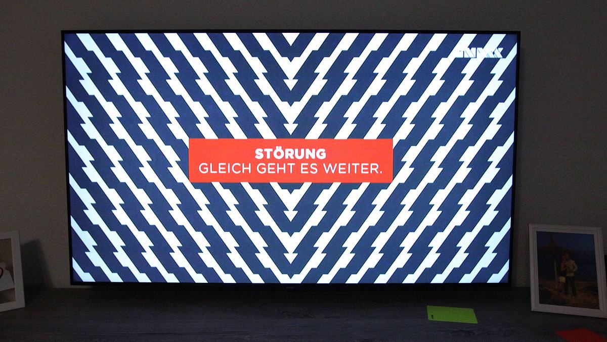 TV Störbild Man sieht einen modernen Fernseher mit einem Störbild. In der Mitte des TVs steht Störung. Gleich geht es weiter: