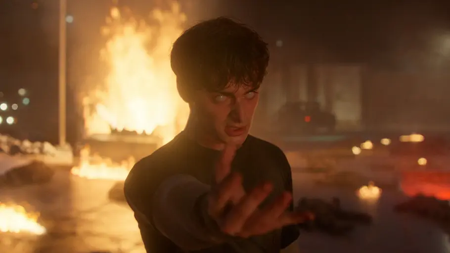 „Stranger Things 5“ Eine Person mit weiß leuchtenden Augen steht in einer dramatischen Szene im Vordergrund und streckt bedrohlich die Hand nach vorne aus. Im Hintergrund lodern große Flammen und Feuerstellen, die Umgebung wirkt zerstört und rauchig, was auf Chaos oder einen Kampf hindeutet.