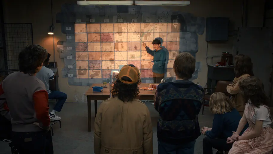 „Stranger Things 5“ Eine Gruppe von sieben Jugendlichen steht und sitzt in einem abgedunkelten Raum. Sie blicken auf eine große Wand, an der ein großes Raster aus Papierblättern angebracht ist, das wie eine riesige Karte aussieht. Einer der Jugendlichen steht vorne und erklärt etwas, wobei er mit einem Zeigestab auf die Karte zeigt.
