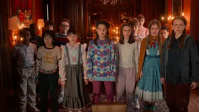 „Stranger Things“: Eine Gruppe von etwa zwölf Kindern steht dicht nebeneinander in einem altmodisch eingerichteten Raum mit Holzwänden, Spiegeln und einem Kronleuchter. Die Kinder blicken ernst und gespannt nach vorne. Sie tragen verschiedene, teils gemusterte Kleidung im Retro-Stil, darunter ein T-Shirt mit der Aufschrift E.T. PHONE HOME. Im Hintergrund stehen Kerzen auf Tischen und Spiegel reflektieren das Licht, wodurch eine geheimnisvolle Stimmung entsteht. - Foto: Netflix