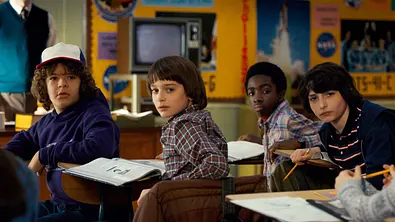 Die vier Hauptfiguren aus Stranger Things sitzen in einem Klassenzimmer und schauen über ihre Schulter nach hinten. - Foto:  IMAGO / Everett Collection