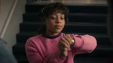 Eine Jugendliche mit lockigen Haaren und rosa Pullover steht vor einer Treppe in einer Schule und schaut prüfend auf ihre Armbanduhr. - Foto: © 2025 Netflix, Inc. Eine Jugendliche mit lockigen Haaren und rosa Pullover steht vor einer Treppe in einer Schule und schaut prüfend auf ihre Armbanduhr. - Foto: © 2025 Netflix, Inc.