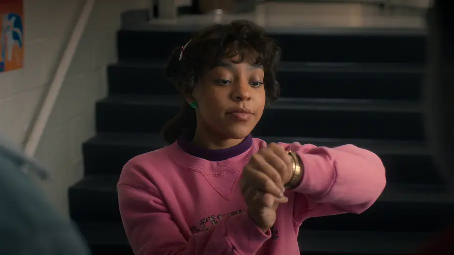„Stranger Things 5“ Eine Jugendliche mit lockigen Haaren und rosa Pullover steht vor einer Treppe in einer Schule und schaut prüfend auf ihre Armbanduhr.