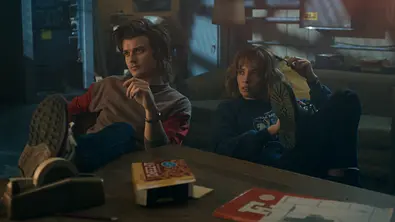 Zwei Jugendliche sitzen entspannt auf Stühlen in einem schummrig beleuchteten Raum und haben die Beine auf einen Tisch gelegt. - Foto: Courtesy of Netflix © 2025