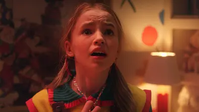 Ein junges Mädchen mit blondem Haar und bunter Kleidung steht in einem Kinderzimmer und blickt erschrocken nach oben. Im Hintergrund sind bunte Wanddekorationen, eine Lampe und ein Kuscheltier zu sehen. - Foto: 2025 Netflix, Inc.