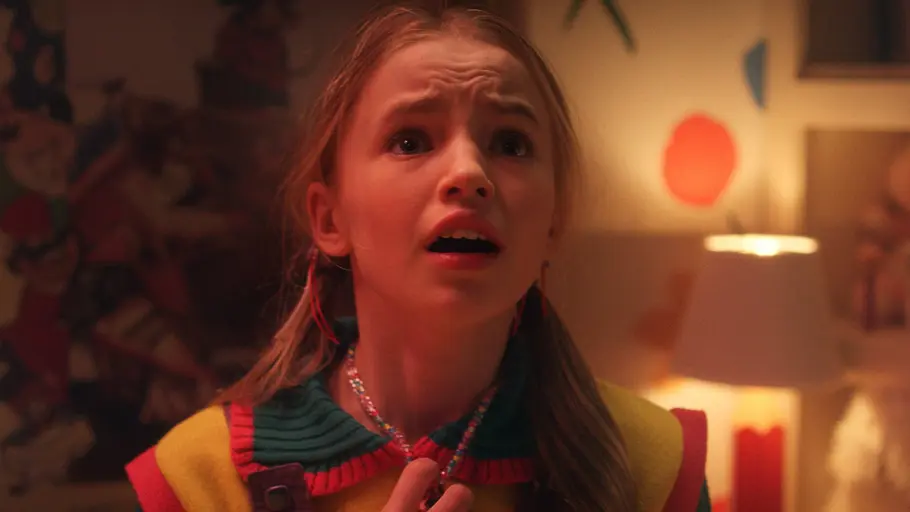 „Stranger Things 5“ Ein junges Mädchen mit blondem Haar und bunter Kleidung steht in einem Kinderzimmer und blickt erschrocken nach oben. Im Hintergrund sind bunte Wanddekorationen, eine Lampe und ein Kuscheltier zu sehen.