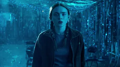 Mädchen steht in dunkler, geschmückter Turnhalle und schaut erschrocken. - Foto: Courtesy of Netflix © 2022