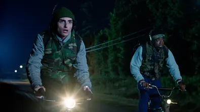 Zwei Jugendliche (Mike und Lucas) fahren nachts auf Fahrrädern eine dunkle Straße entlang. Beide tragen Camouflage-Westen und Rucksäcke; der Vorderste hat eine grüne Mütze auf, der andere ein Stirnband. Die Szene ist von Spannung und Eile geprägt. - Foto: Courtesy of Netflix © 2025