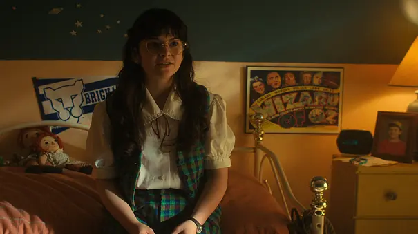 Ein Mädchen mit langen braunen Haaren und Brille sitzt auf einem Bett in einem gemütlich eingerichteten Jugendzimmer. Das Zimmer ist im warmen Licht einer Nachttischlampe beleuchtet. - Foto: Courtesy of Netflix © 2022
