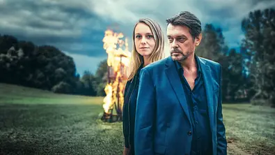 Hary Prinz steht vor Miriam Unterberger. Hinter ihnen ist auf einem Feld ein brennender Hochsitz.  - Foto: ORF/ARD Degeto Film/Allegro Film/Stefan Robitsch