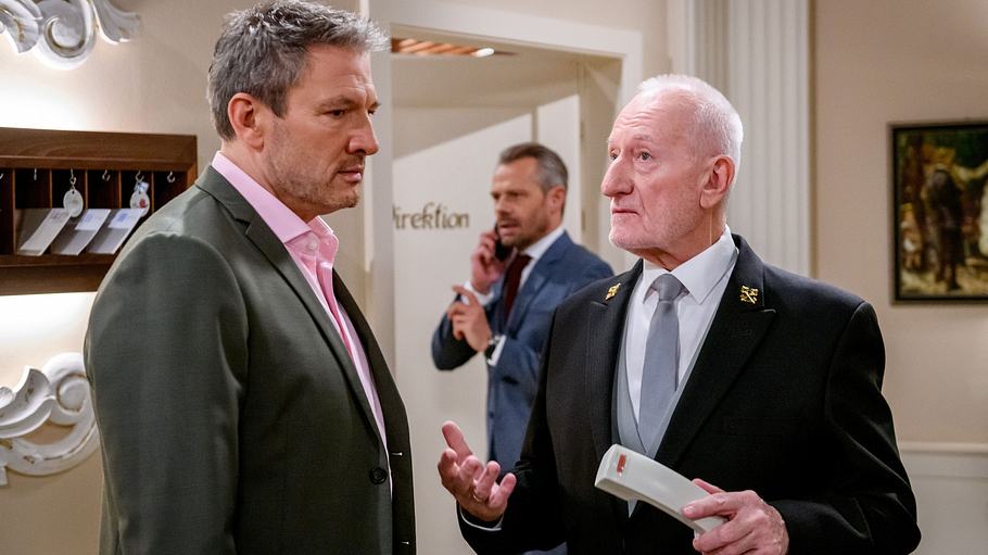 Christoph und Alfons stehen am Fürstenhof zusammen. Alfons hält den Telefonhörer in der linken Hand, Christoph schaut leer ins Weite. Erik ist telefoniert im Bildhintergrund zu sehen.