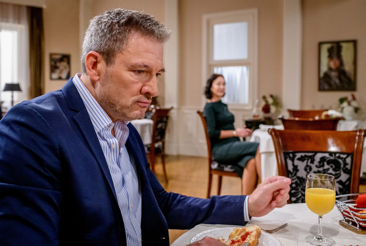 Christoph und Sophia bei Sturm der Liebe Christoph starrt gedankenverloren nach vorne während er am Fürstenhof isst. Sophia sitzt verschwommen im Hintergrund und sieht ihn hoffnungsvoll an.