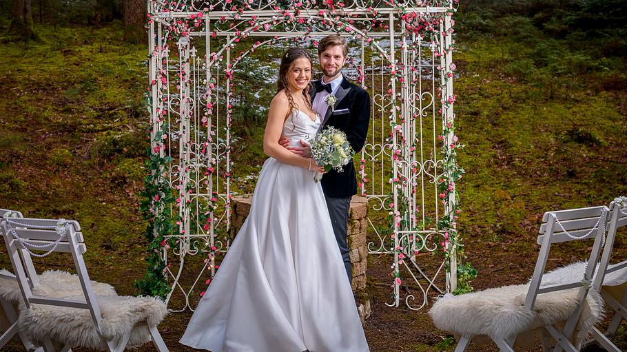 Maxi und Henry sind im Hochzeitskleid bzw. Anzug zu sehen. Sie stehen vor dem Wunschbrunnen und schauen glücklich in die Kamera