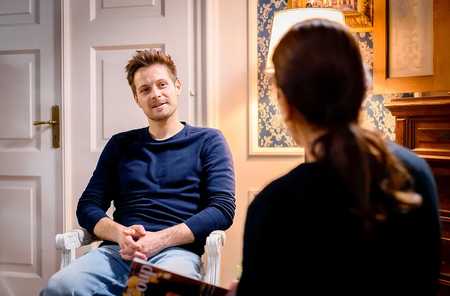 Sturm der Liebe-Vorschau: Adam (Philip Birnstiel) sitzt mit gefalteten Händen Mona (Daniela Ziegler) gegenüber, die von hinten zu sehen ist.
