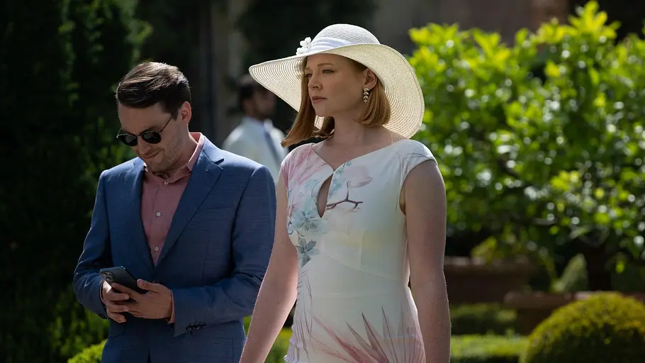 Succession Roman Roy blickt auf sein Smartphone, während Shiv Roy in einem weißen Kleid mit großem Sonnenhut entschlossen nach vorn schaut – eine Szene aus der Dramaserie „Succession“.