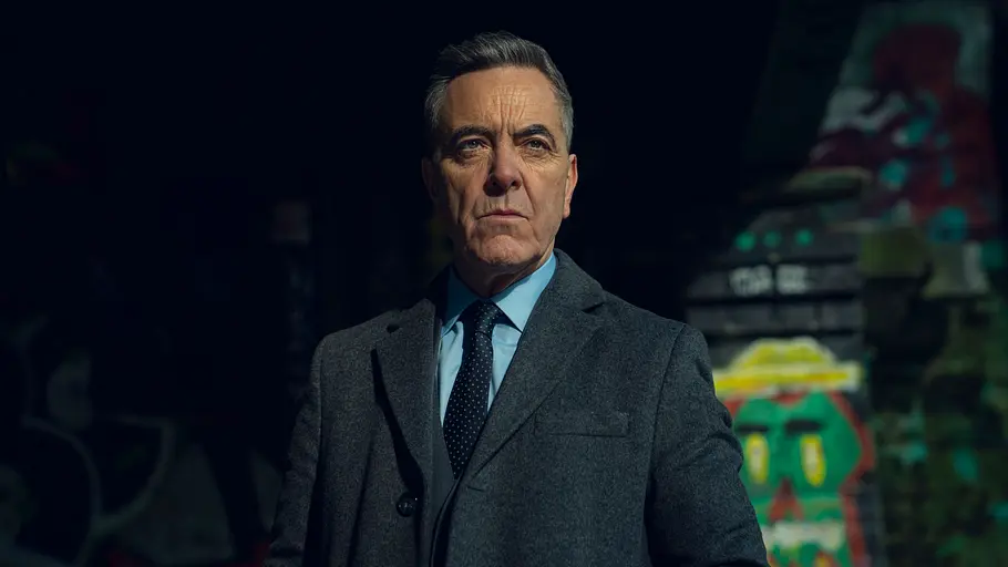 James Nesbitt Netflix Schauspieler James Nesbitt steht in Mantel und Krawatte vor einer Backsteinwand und schaut in die Ferne