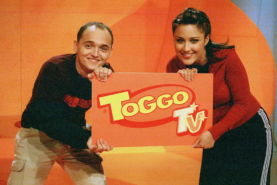 Nina Moghaddam und Paddy Kroetz halten gemeinsam ein Schild mit dem Logo von „Toggo TV“ in die Kamera. Die beiden Moderator:innen lächeln in einem orangefarbenen TV-Studio und präsentieren die Kindersendung.