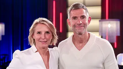 Susan Link und Micky Beisenherz stehen lächelnd in weißen Oberteilen nebeneinander vor blau und rot beleuchtetem Hintergrund. - Foto: IMAGO / Future Image