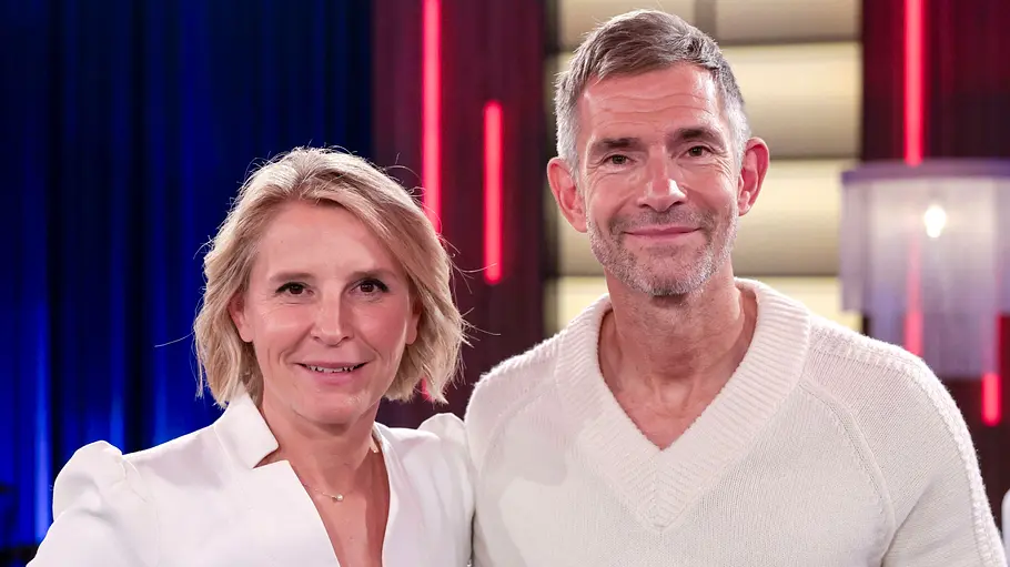 Susan Link und Micky Beisenherz Susan Link und Micky Beisenherz stehen lächelnd in weißen Oberteilen nebeneinander vor blau und rot beleuchtetem Hintergrund.