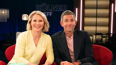 Susan Link und Micky Beisenherz sitzen nebeneinander im Studio der Talkshow „Kölner Treff“ und blicken lächelnd in die Kamera. - Foto: WDR/Melanie Grande