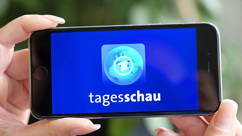 Person hält in Handy in beiden Händen und sieht darauf das Logo der Tagesschau. - Foto:  IMAGO / epd