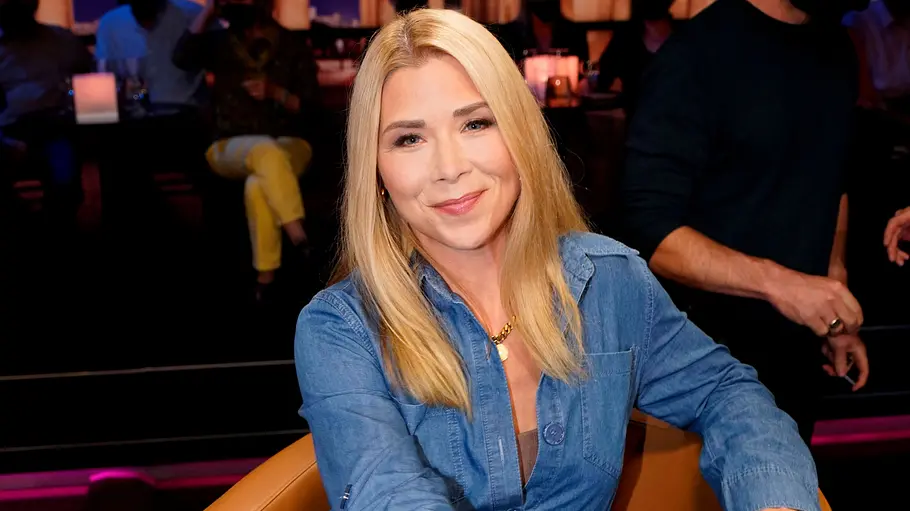 Tanja Szewczenko Umzug Rumänien. Tanja Szewczenko im blauen Jeanshemd sitzt lächelnd in einem braunen Sessel. Im Hintergrund sind weitere Personen zu sehen.