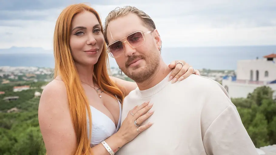 Tara Tabitha und Dennis Lodi haben Höhen und Tiefen hinter sich. Tara mit langen roten Haaren und Dennis mit Sonnenbrille posieren eng beieinander vor grüner Küstenlandschaft und Meer.