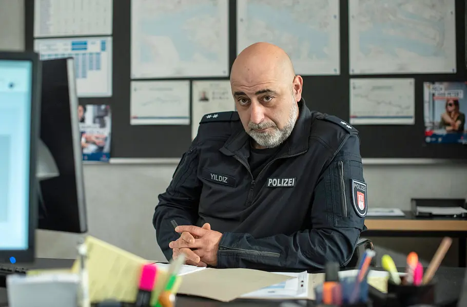 Polizeibeamter in Uniform sitzt an einem Schreibtisch in einem Büro und blickt ernst in die Kamera, im Hintergrund Karten und Einsatzunterlagen an der Wand.