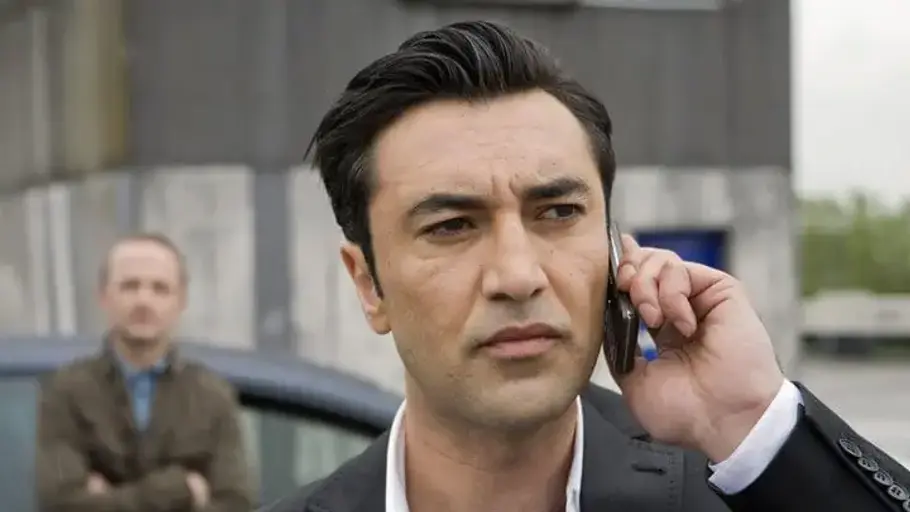 Tatort Mehmet Kurtulus steht mit einem Handy an dem Ohr vor einem Mann, der an einem Auto lehnt.