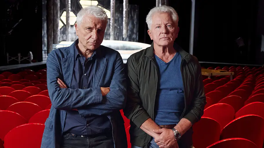 Miroslav Nemec (l.) und Udo Wachtveitl stehen in einem Theater. Die Stühle hinter ihnen sind rot. 
