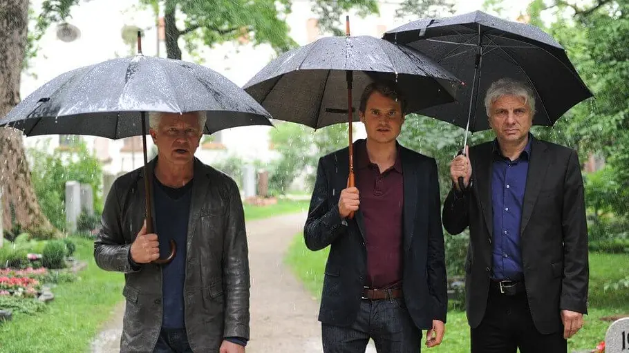 Batic, Engelhardt und Leitmayr stehen mit Regenschirmen im Regen.