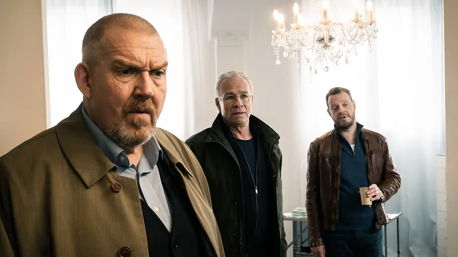 Tatort - Diesmal ist es anders Dietmar Bär und Klaus J. Behrendt stehen mit einer Assistenzkraft in einem weißgeschmückten Saal. An der Decke ist ein Kronleuchter.