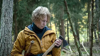 Corinna Harfouch steht mit einem gelben Mantel im Wald. In ihrer Hand hält sie eine Axt.  - Foto: rbb/Conny Klein