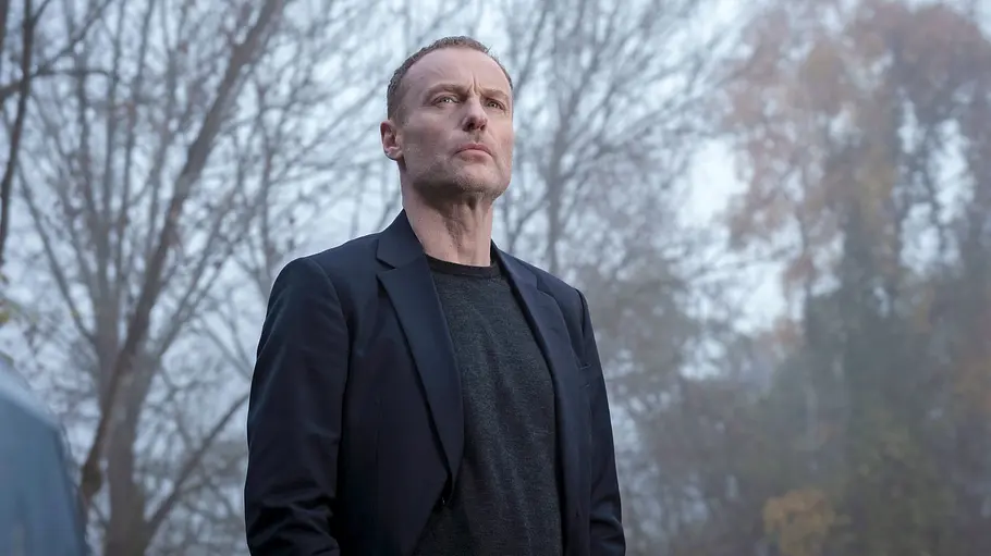 Mark Waschke steht mit schwarzem Mantel im Wald.