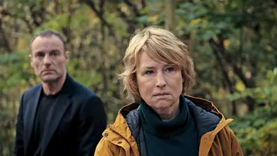 Mark Waschke (l.) und Corinna Harfouch stehen im Wald. Harfouch trägt eine gelbe Jacke, Waschke einen schwarzen Mantel. - Foto: rbb/Conny Klein