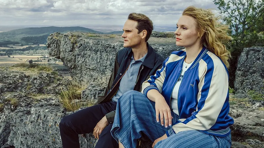Tatort: Gottesgarten Voss und Rathgeber in blau-weißer Jacke sitzen auf Felsen vor einer weiten Hügellandschaft unter bewölktem Himmel.