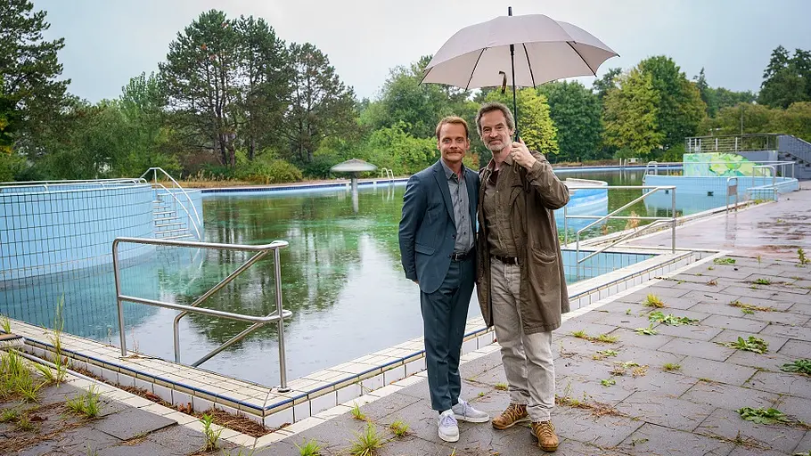 Faber und Kossik stehen im Regenwetter vor einem Schwimmbecken.