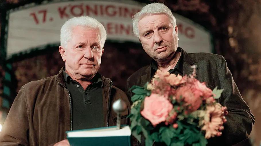 Tatort - Königinnen Miroslav Nemec und Udo Wachtveitl stehen für den Münchner „Tatort: Königinnen“ mit Blumen vor einem Banner.