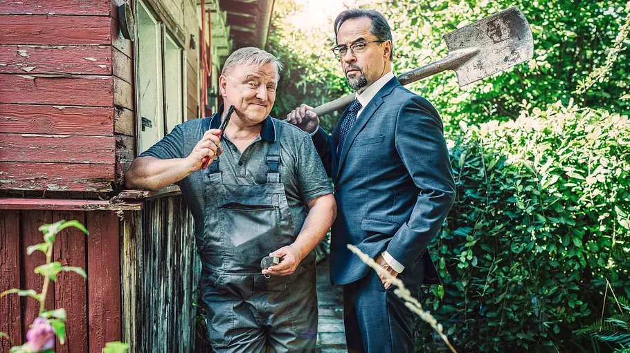 Tatort: Unter Gärtnern Axel Prahl steht im Blaumann und Jan Josef Liefers im Anzug in einem Schrebergarten. Beide halten Werkzeuge in der Hand.
