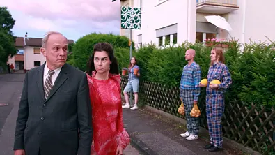 Ulrich Tukur (l.) steht neben mehreren Personen in einer Fantasieverison einer Wohngegend.  - Foto: HR/Senator Film/Dietrich Brüggemann