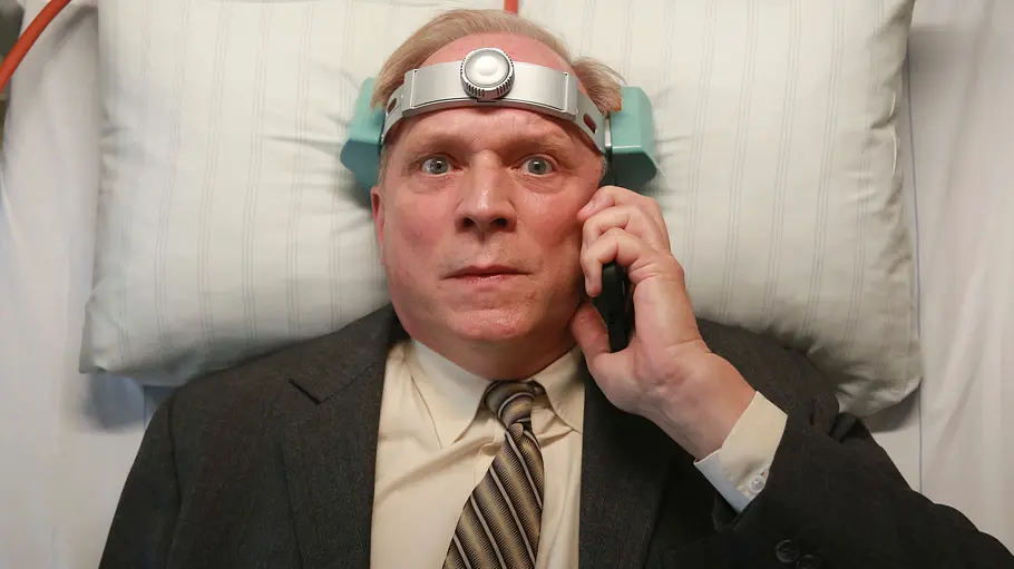 Ulrich Tukur liegt mit einer Apparatur um den Kopf auf einem Krankenhausbett. In seiner Hand ist ein Handy.