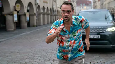 Jan Josef Liefers rennt in einem Hawaii-Hemd vor einem Auto weg. - Foto: MDR/WDR/Bavaria Fiction GmbH/Thomas Kost