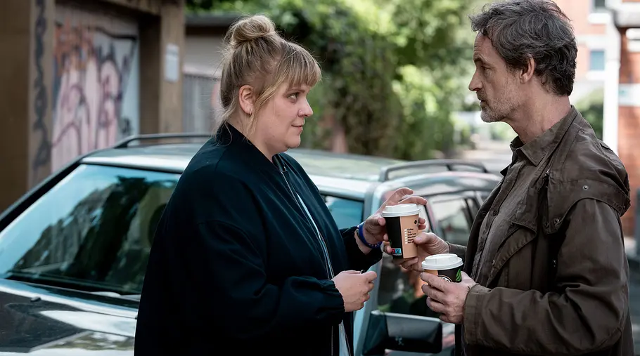 Tatort – Schmerz Stefanie Reinsperger (l.) steht neben Jörg Hartmann. Er hat Kaffee in der Hand. Im Hintergrund sieht man ein Auto.