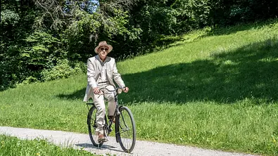 Ermittler Murot fährt mit einem Fahrrad an einer grünen Wiese vorbei. - Foto: HR/Bettina Müller